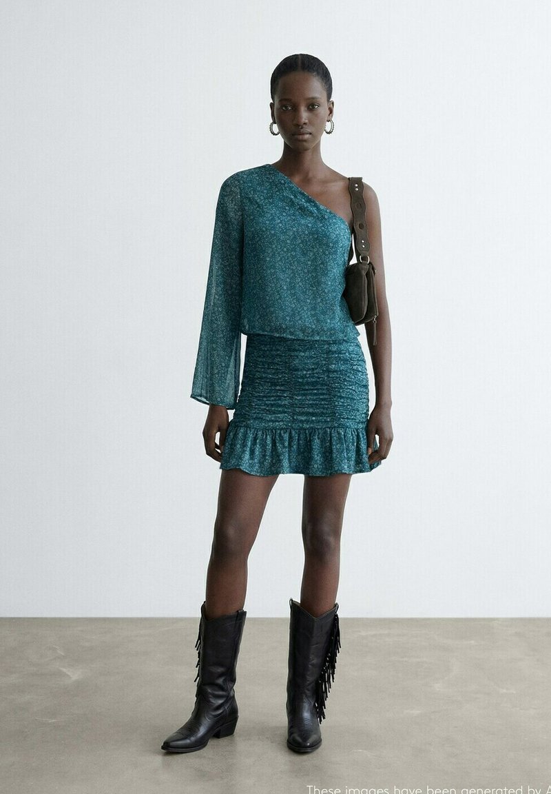 Femme portant une robe asymétrique bleu sarcelle à ourlet volanté, bottes noires à franges, boucles d'oreilles créoles et sac à bandoulière marron, debout sur un sol gris.