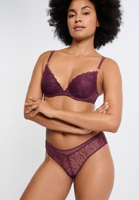 Reggiseno a triangolo in pizzo viola scuro con motivo decorativo a foglia e sottili spalline regolabili; perizoma trasparente coordinato mostrato sotto.