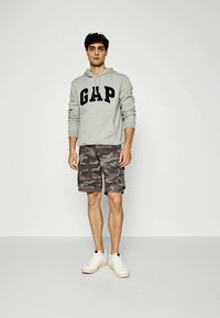 Grey hoodie with "GAP" logo, paired with gray camo shorts, standing on a neutral background. White sneakers complete the casual outfit.