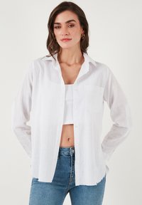 White linen button-up shirt with a pointed collar, single pocket, and frayed hem, worn over a white crop top and paired with blue jeans.