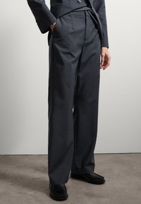Pantalons gris foncé ajustés avec une taille haute, des jambes larges et des poches latérales. Associés à des mocassins noirs, avec un design texturé.