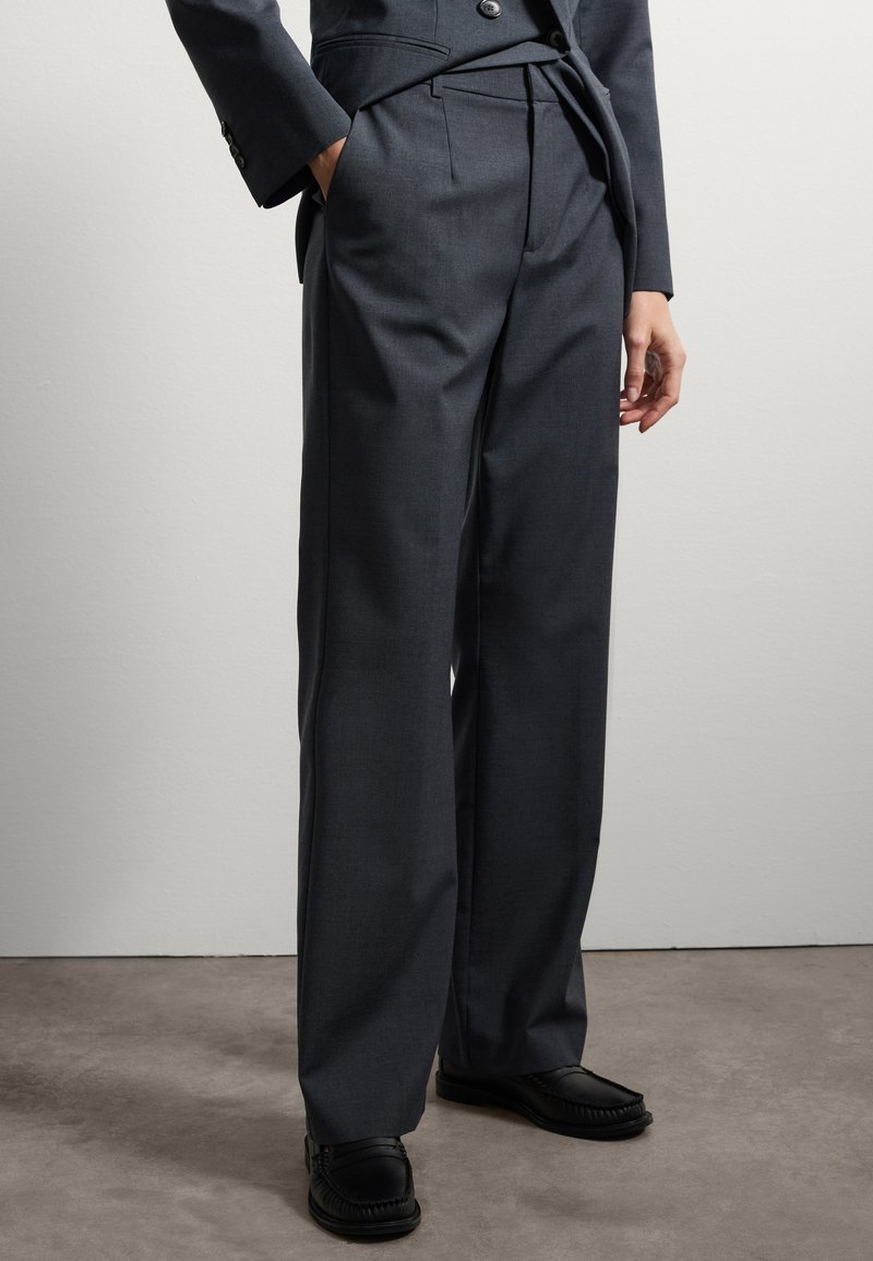 Pantaloni tailorati grigio scuro con vita alta, gambe larghe e tasche laterali. Abbinati a mocassini neri con un design testurizzato.