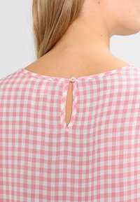 Vista posteriore di una persona che indossa una blusa a scacchi rosa e bianca con una piccola chiusura a bottone alla nuca e un'apertura sotto di essa.