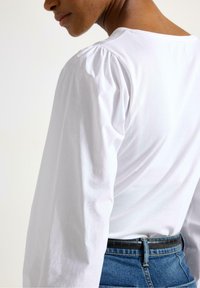 Una maglietta bianca a maniche lunghe presenta spalle a sbuffo, un collo rotondo e un tessuto morbido e liscio. Abbinata a jeans blu a vita alta e una cintura nera.