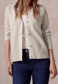 Žena v svetle béžovém cardiganu na knoflíky přes bílé tričko a tmavě modré džíny, stojící proti neutrálnímu pozadí.
