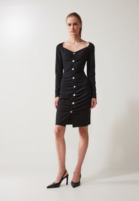 Robe noire à manches longues avec décolleté cœur, détail froncé et une rangée de boutons argentés. Présente une fente haute cuisse et un tissu doux.