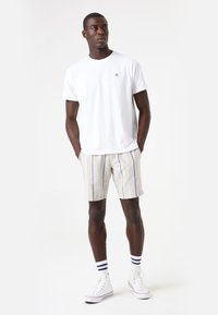 Camiseta de algodón blanca con un pequeño logo en el pecho, combinada con pantalones cortos rayados beige, zapatillas deportivas blancas y calcetines a rayas azules.