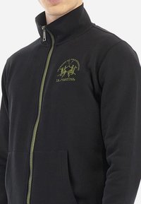 Černá bunda na zip z hladké tkaniny s vysokým límcem, olivově zeleným vyšívaným logem a přední kapsou.