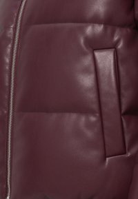 Veste matelassée en cuir bordeaux avec fermeture éclair argentée et détail de poche rectangulaire. Texture lisse et design matelassé avec rembourrage.