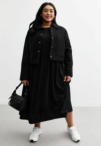 Veste noire cropped en tissu texturé avec des boutons-pression, superposée sur une longue robe noire. Les accessoires comprennent un petit sac à main noir et des baskets blanches.