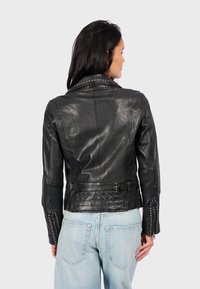 Chaqueta de cuero negra con collar y puños con tachuelas, diseño ajustado, cintura ceñida y textura brillante, llevada sobre jeans de un azul claro.