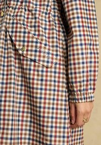 Geruit lang mouw kledingstuk in beige, bruin, rood en marineblauw met een borstzak met knopen en elastische manchetten, gedragen door een persoon met een gouden ring aan de vinger.