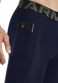 Marinefarbene Sporthosen mit einem breiten schwarzen Bund und einer Seitentasche, in der ein Smartphone gehalten wird. Glattes, dehnbares Material mit sichtbaren Nähten.