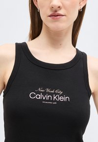 Νεαρή γυναίκα που φοράει μαύρο ριμπ αμάνικο τοπ με το κέντημα "New York City Calvin Klein Established 1968" στο στήθος.