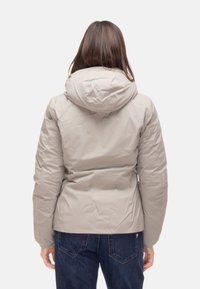 Giacca isolata grigio chiaro con cappuccio, caratterizzata da una texture liscia e una vestibilità su misura, indossata sopra jeans blu scuro.