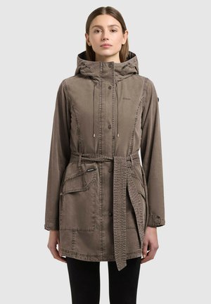 Kobieta w brązowym płaszczu z kapturem, z przednimi kieszeniami i wiązanym paskiem, stojąca na tle jasnego, jednolitego tła.