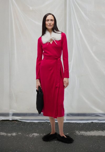 Rochie roșie tip wrap cu guler alb din blană, mâneci lungi, talie legată și material neted. Poartă o geantă neagră împletită în mână și papuci negri pufoși.