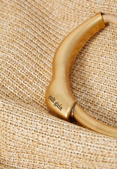 A gold-toned bangle with "cult gaia" engraved rests on a light, woven straw texture.
