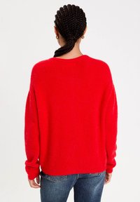 Kvinde med fletninger, bundet tilbage, iført guldøreringe, rød fuzzy sweater og blå jeans, der vender væk på hvid baggrund.