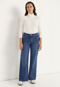 Blauwe wijde denim jeans met een hoge taille, voorzien van een sluiting met een knoop aan de voorkant en twee zijzakken. Gecombineerd met een witte coltrui.