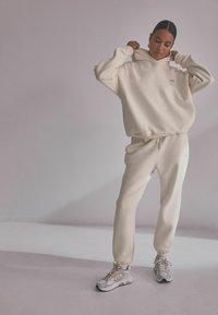 Cream-colored oversized hoodie and matching joggers made of fleece. Features a drawstring at the waist and ribbed cuffs. Paired with gray sneakers.