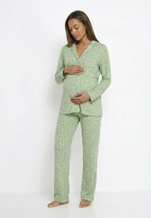 Femme enceinte en pyjama vert clair à imprimé léopard, debout pieds nus, tenant doucement son ventre avec les deux mains sur fond blanc.