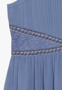 Jasnoniebieska sukienka z plisowanym materiałem, z teksturowanym panelem koronkowym i ozdobnymi koralikami ułożonymi diagonalnie wzdłuż talii.