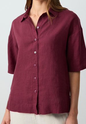 Femme portant une chemise à manches courtes, boutonnée, avec col, en tissu rouge foncé, associée à un pantalon clair.