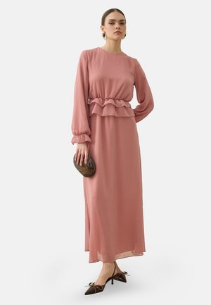 Donna che indossa un lungo vestito rosa con vita e maniche arruffate, tiene una pochette ovale scura e indossa tacchi a punta marroni con fiocchi.