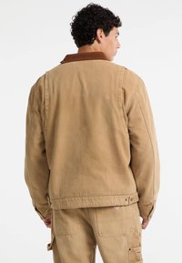 Giacca in tela color sabbia con colletto in corduroy marrone, cuciture visibili e dettagli delle tasche laterali. Indossata con pantaloni cargo abbinati.