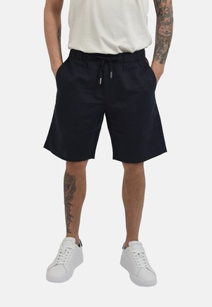Uomo che indossa pantaloncini neri fino al ginocchio, sneaker bianchi e una camicia bianca con braccia e gambe tatuate visibili.