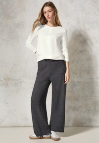 Weißes Langarmoberteil mit einer Brustnaht, kombiniert mit grauen weit geschnittenen Hosen mit Taschen und weißen Sneakers mit hellbraunen Sohlen.