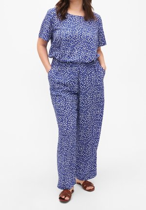 Vrouw draagt een blauw-wit gestippeld shirt met korte mouwen en bijpassende wijde broekspijpen, met bruine sandalen met open tenen, handen in de zakken.