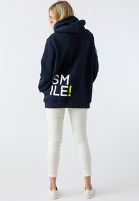 Felpa navy con un grande grafiche bianco "SMILE!" e dettagli in verde neon, abbinata a pantaloni bianchi aderenti e scarpe bianche casual.