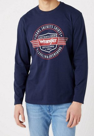 Mężczyzna w granatowej, długorękawowej koszuli Wrangler z czerwoną i białą okrągłą logo oraz jasnoniebieskich dżinsach, stojący na tle jednolitego tła.