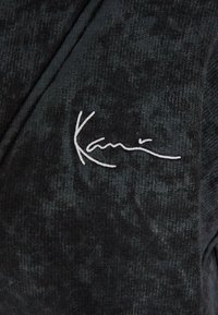 Juodas tekstūruotas marškiniai su subtiliu raštu, puošti balta siuvinėta "Kani" logo ant krūtinės.