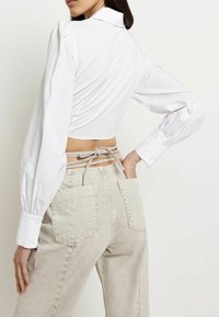 Camisa blanca corta con mangas largas abullonadas, que revela un detalle de lazo en la parte posterior. Combinada con unos jeans de tiro alto en un tono beige claro que cuentan con bolsillos traseros.