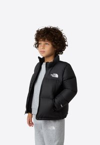 Svart dunjakke med høy krage og logo på brystet, brukt over en grå genser, kombinert med grå joggebukser. Krøllete hår er synlig.