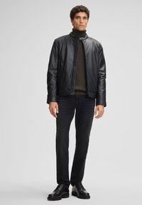 Giacca in pelle nera con colletto alto, indossata sopra un maglione verde scuro, abbinata a jeans neri aderenti e scarpe stringate nere.