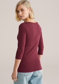 Bordowa bluzka z rękawem do połowy przedramienia wykonana z gładkiej tkaniny, z zaokrąglonym dekoltem i dopasowanym krojem. Połączona z jasnoniebieskimi dżinsami.