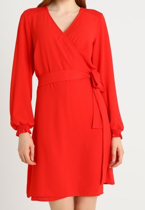 Femme portant une robe portefeuille rouge à manches longues avec ceinture à nouer et poignets froncés, vue du cou jusqu'à la mi-cuisse sur fond blanc.
