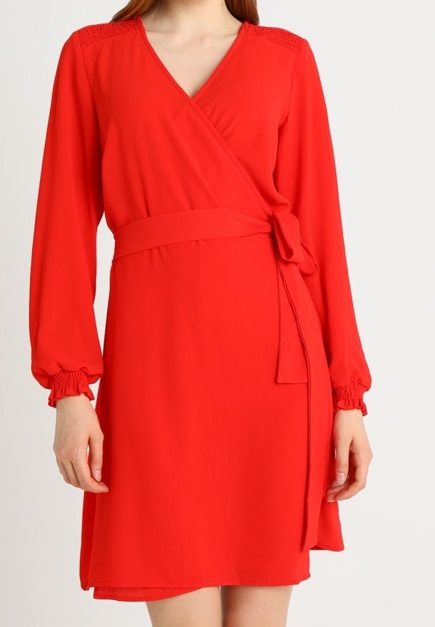 Femme portant une robe portefeuille rouge à manches longues avec ceinture à nouer et poignets froncés, vue du cou jusqu'à la mi-cuisse sur fond blanc.