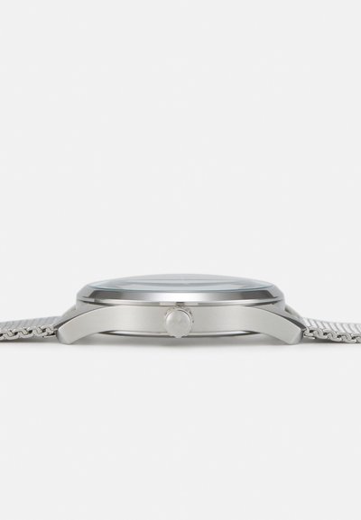 Montre en acier inoxydable argenté avec un boîtier rond, un bracelet en maille et une couronne proéminente sur le côté. Le verre transparent révèle un cadran simple.