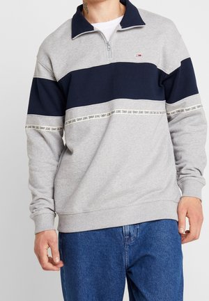 Man draagt een grijze en marineblauwe quarter-zip trui met "Tommy Jeans" tekstbanden en blauwe spijkerbroek, staand tegen een effen achtergrond.