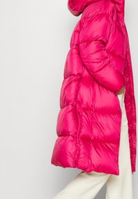 Abrigo acolchado rosa con acabado brillante, que cuenta con capucha, secciones acolchadas de gran tamaño y cremallera frontal. Combinado con pantalones de color claro.