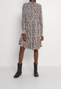 Blumenkleid mit langen Ärmeln und einem geschichteten Saum. Die Farben umfassen Lila, Gelb und Grün auf schwarzem Hintergrund. Kombiniert mit schwarzen Stiefeln.
