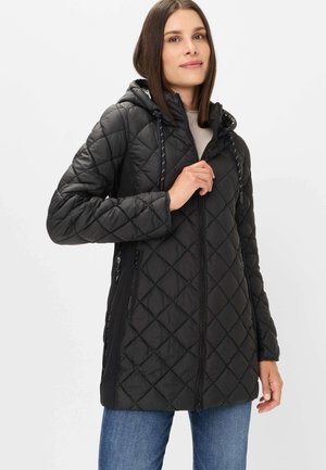 Frau in einer schwarzen gesteppten Kapuzenjacke mit Reißverschluss, die den Reißverschluss anpasst, trägt einen hellen Pullover und blaue Jeans.