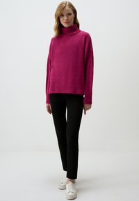 Pull col roulé magenta en tissu doux et texturé, associé à un pantalon noir et des baskets blanches. Coupe décontractée avec des accents en maille.