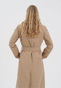 Femme aux longs cheveux blonds ondulés, portant un trench-coat beige ceinturé, debout les mains sur les hanches, dos tourné sur un fond uni.