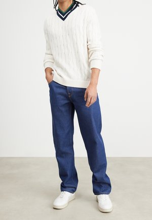 Mies, jolla on valkoinen kaapelineuleinen V-kaulusneule, siniset farkut ja valkoiset lenkkitossut, seisoo yhdellä kädellä taskussa yksiväristä taustaa vasten.